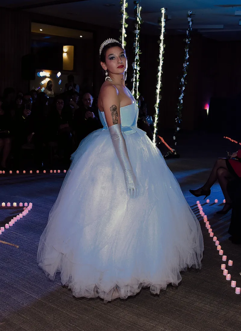 Modelo en pasarela interior usando un vestido celeste claro con corpiño strapless, cintura marcada y falda muy amplia de tul; lo combina con guantes largos y tiara.