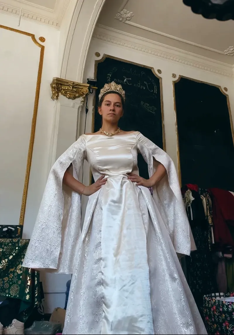 Persona de pie con vestido marfil satinado de falda larga, mangas abiertas tipo capa, textura brocada y corona dorada en un interior ornamentado.