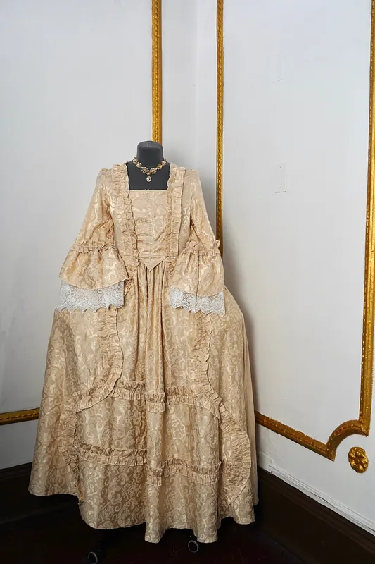 Vista frontal del vestido Charlotta en maniquí, con escote cuadrado, cuerpo ajustado, mangas con encaje blanco, falda amplia y vuelos fruncidos sobre brocado color champaña.