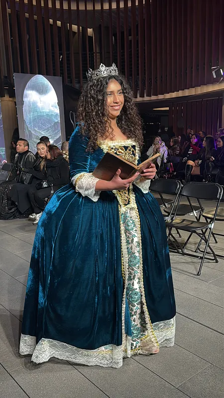 Persona de pie con vestido azul petróleo de falda amplia, corpiño dorado, encaje claro, corona plateada y libro abierto en las manos.